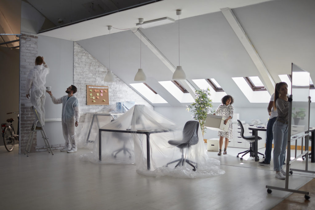 Young entrepreneurs moving in the office. Men painting brick wall. Other coworkers moving office equipment and renovating their office.